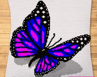 Op de afbeelding: Een vierkant wit gehaakt kussen met een gepixeld vlinderontwerp in levendige tinten blauw, paars en zwart. De vlinder heeft witte stippen op de zwarte vleugelranden. Een klein roze vakje bevat de tekst: SYMBOLIC IMAGE, NOT A PHOTO!