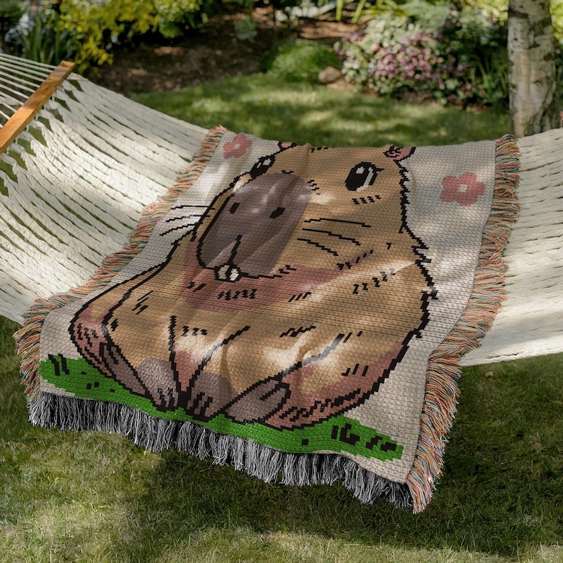 Puede incluir: Una manta de tejido de tapicer&iacute;a con un capibara de dibujos animados con pelaje marr&oacute;n, nariz rosa y ojos negros. La manta tiene un fondo beige con flores rosas.