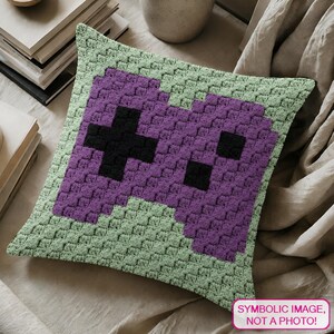 May include: A square, crocheted pillow featuring a pixelated video game controller design. The controller is purple with a black cross and black square buttons, set against a light green border. The pillow is on a neutral-toned surface.