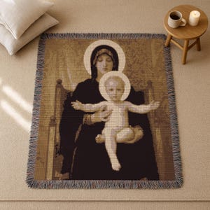 May include: A sepia-toned woven throw blanket featuring a religious image of a woman holding a child, both with halos. The blanket has a fringed edge and is placed on a neutral-colored surface. The image depicts a classic religious theme.