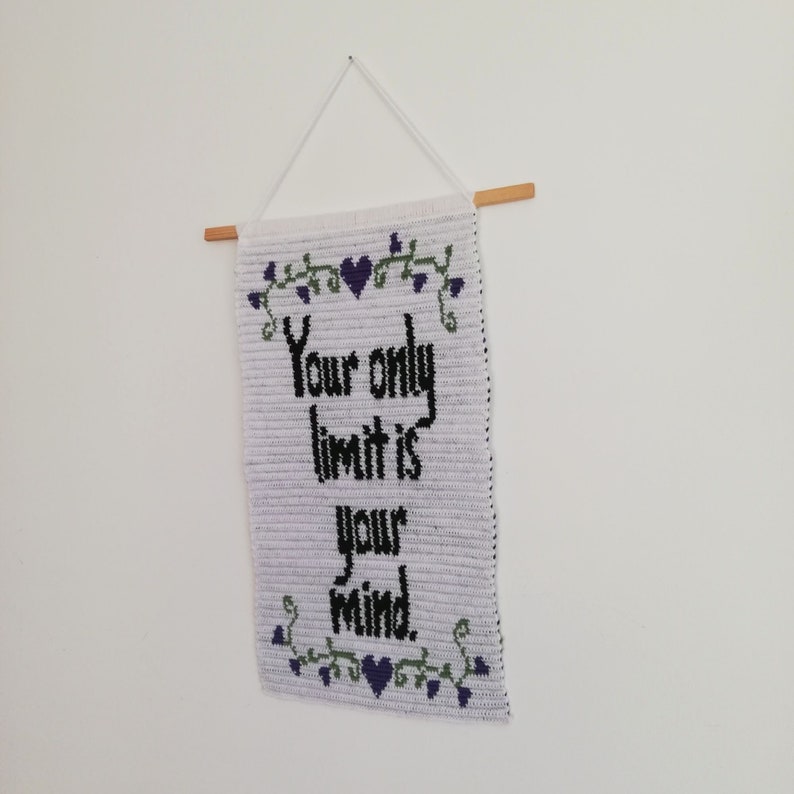 Pu&ograve; includere: Un arazzo da parete in crochet bianco con il testo "Your only limit is your mind" in lettere nere. L'arazzo &egrave; decorato con viti verdi e fiori viola.