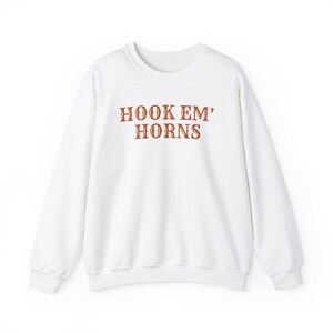 Könnte beinhalten: Weißes Sweatshirt mit Rundhalsausschnitt und dem Text "HOOK EM' HORNS" in einem gebrannten Orange. Das Sweatshirt besteht aus einem weichen Material und hat lange Ärmel. Ein lässiges und bequemes Oberteil.