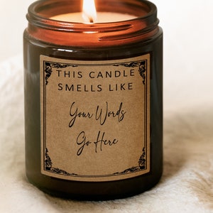 May include: A lit brown glass candle jar with a label that reads "THIS CANDLE SMELLS LIKE Your Words Go Here." A hand holds a lit match above the candle. The candle is on a white surface.