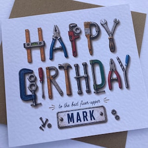 May include: A square greeting card featuring the words HAPPY BIRTHDAY formed by illustrations of various hand tools including wrenches, pliers, and hammers. Below this, the text reads to the best fixer-upper and MARK on a metal plate, with small screws.