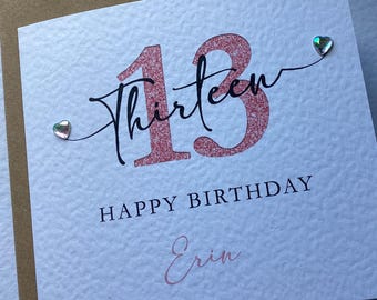 May include: A square white greeting card with a textured surface featuring a large rose gold glitter number 13. The word thirteen is written in black script across the number. Below is the text HAPPY BIRTHDAY and the name Erin in rose gold.