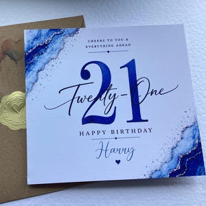 May include: A square birthday card with a blue and silver marble-style border, featuring the text 21 Twenty-One HAPPY BIRTHDAY Harry, CHEERS TO YOU & EVERYTHING AHEAD, and a small heart icon, paired with a brown envelope sealed with a gold sticker.