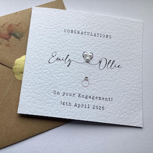 May include: A square white textured greeting card with the text CONGRATULATIONS, Emily and Ollie, On your Engagement! 14th April 2026, featuring a heart with three white pearls and a small ring graphic, paired with a brown envelope with a gold seal.