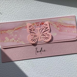 K&ouml;nnte beinhalten: Rosa Karte mit einem rosa Glitzer-Schmetterlingsausschnitt und dem Namen "Indie" in schwarzer Kursivschrift.