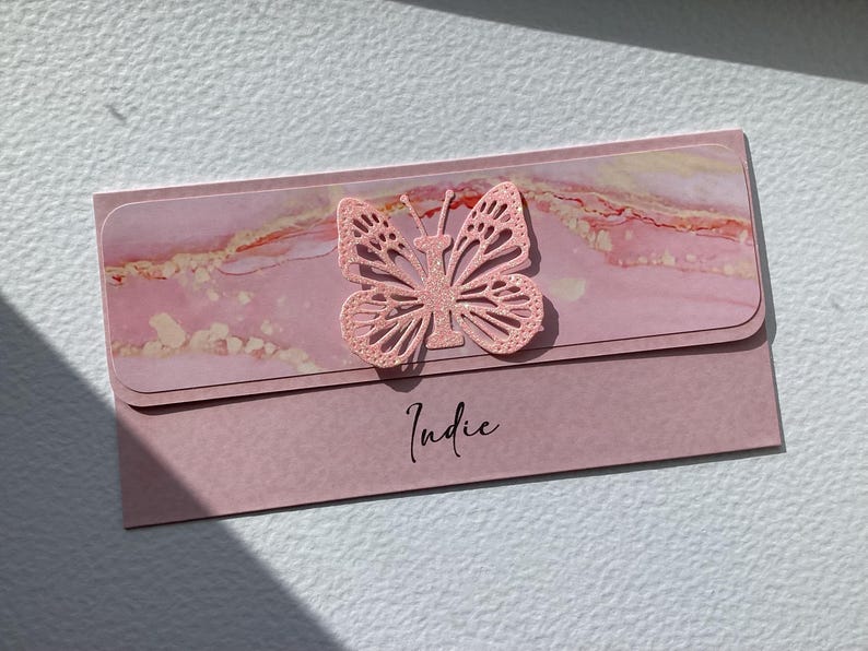 K&ouml;nnte beinhalten: Rosa Karte mit einem rosa Glitzer-Schmetterlingsausschnitt und dem Namen "Indie" in schwarzer Kursivschrift.