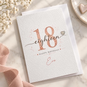 May include: A white textured greeting card featuring the number 18 in rose gold glitter, the word eighteen in black script, and the text Happy Birthday Eve. The design includes small heart accents, a silver heart embellishment, and a small crystal.
