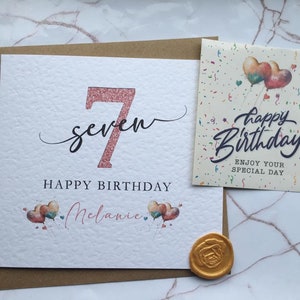May include: A pink glitter 7 with the word "seven" in cursive script. The card says "Happy Birthday Melanie" with a pink and blue heart balloon design. The card also includes a separate card with the text "Happy Birthday" and "Enjoy your special day" with a colorful balloon design.
