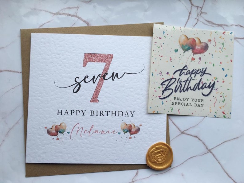 May include: A pink glitter 7 with the word "seven" in cursive script. The card says "Happy Birthday Melanie" with a pink and blue heart balloon design. The card also includes a separate card with the text "Happy Birthday" and "Enjoy your special day" with a colorful balloon design.
