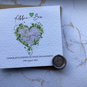 May include: A white card with a green heart-shaped map design and the text "Abbie & Ben" and "Congratulations on your engagement 29th August 2022". A brown envelope and a heart-shaped wax seal are in the background.
