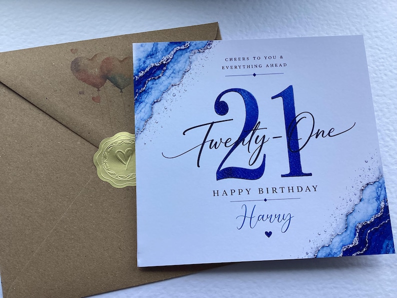 May include: A square birthday card with a blue and silver marble-style border, featuring the text 21 Twenty-One HAPPY BIRTHDAY Harry, CHEERS TO YOU & EVERYTHING AHEAD, and a small heart icon, paired with a brown envelope sealed with a gold sticker.