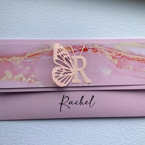 May include: Pink and gold marble patterned card with a pink butterfly and the letter "R" in the center. The name "Rachel" is printed in black at the bottom of the card.