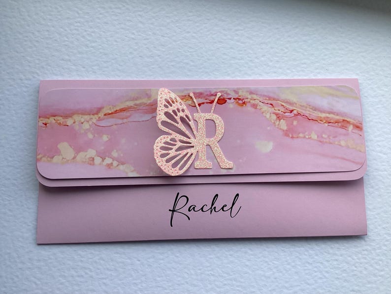 May include: Pink and gold marble patterned card with a pink butterfly and the letter "R" in the center. The name "Rachel" is printed in black at the bottom of the card.