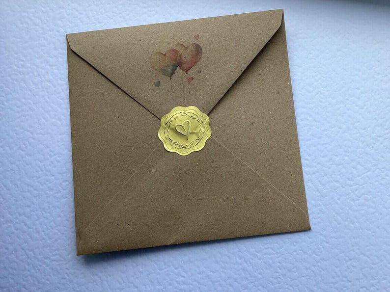 May include: A square brown kraft paper envelope featuring a small illustration of two heart-shaped balloons near the top flap. The envelope is sealed in the center with a round, scalloped-edge gold foil sticker embossed with a heart design.
