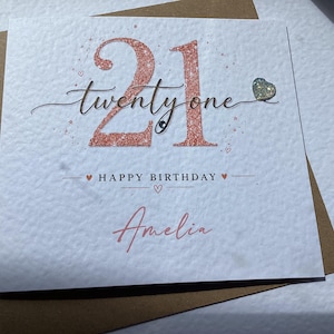 May include: A square white greeting card with a textured surface featuring the rose gold glitter number 21 and the script text twenty one. Below, it reads HAPPY BIRTHDAY with the name Amelia in rose gold script, accented by a small glitter heart.