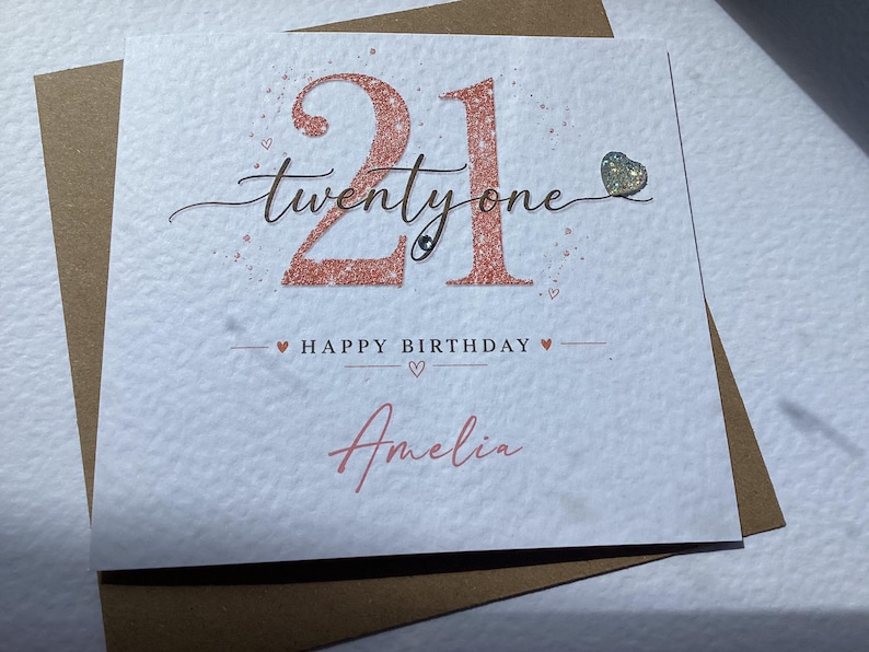 May include: A square white greeting card with a textured surface featuring the rose gold glitter number 21 and the script text twenty one. Below, it reads HAPPY BIRTHDAY with the name Amelia in rose gold script, accented by a small glitter heart.