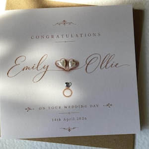May include: A square white greeting card with copper-coloured text reading CONGRATULATIONS, Emily and Ollie, ON YOUR WEDDING DAY, and 14th April 2026. The card features two small heart-shaped embellishments and a ring icon with a heart-shaped stone.