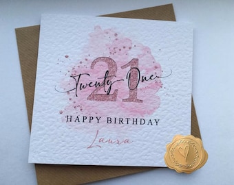 May include: A pink and white card with a watercolor design and the number 21 in gold glitter. The text "Twenty-One" is written in cursive above the number. The text "Happy Birthday Laura" is written below the number.