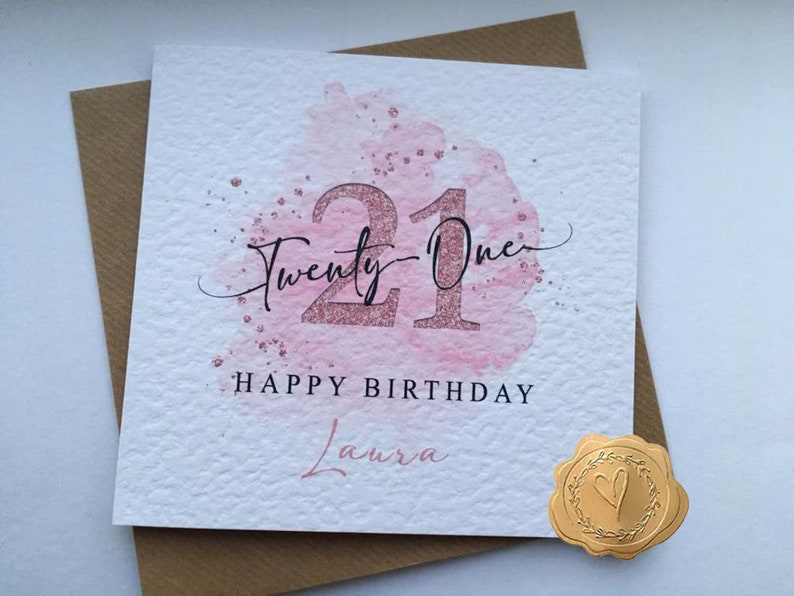 K&ouml;nnte beinhalten: Eine rosa und wei&szlig;e Karte mit einem Aquarelldesign und der Zahl 21 in goldenen Glitzer. Der Text "Twenty-One" ist in Kursivschrift &uuml;ber der Zahl geschrieben. Der Text "Happy Birthday Laura" ist unter der Zahl geschrieben.