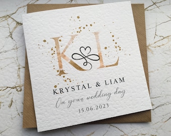 May include: A wedding card with gold foil accents and the initials "KL" in a script font. The names "Krystal & Liam" and the date "15.06.2023" are printed below the initials.