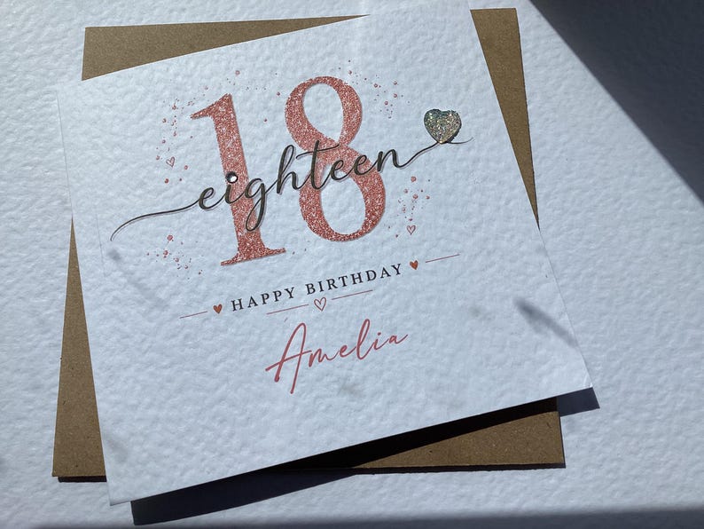 May include: A square white greeting card featuring the large rose gold glitter number 18, the cursive word eighteen, and the text HAPPY BIRTHDAY Amelia in black and rose gold. A small silver heart embellishment is attached to the card surface.