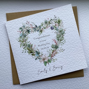 May include: A white wedding card with a green and pink floral heart design. The card says "Congratulations on your Wedding Day 24.09.24" and "Emily & Barry".