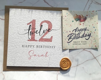 May include: A pink glitter card with the number 12 and the word "twelve" written in cursive. The card says "Happy Birthday Sarah" and has a gold wax seal. A smaller card with confetti and balloons says "Happy Birthday" and "Enjoy your special day."