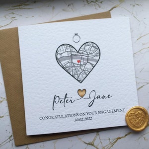 May include: A white card with a black and white map of a city in the shape of a heart. The text "Peter & Jane" is written in black cursive script. The text "Congratulations on your engagement 30.02.2022" is written in black below the heart. A gold wax seal with a heart design is in the bottom right corner.