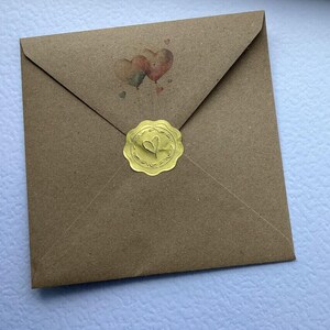 May include: A square brown kraft paper envelope featuring a small illustration of two heart-shaped balloons above the center. The envelope is sealed with a round, scalloped-edge gold foil sticker embossed with a central heart design.