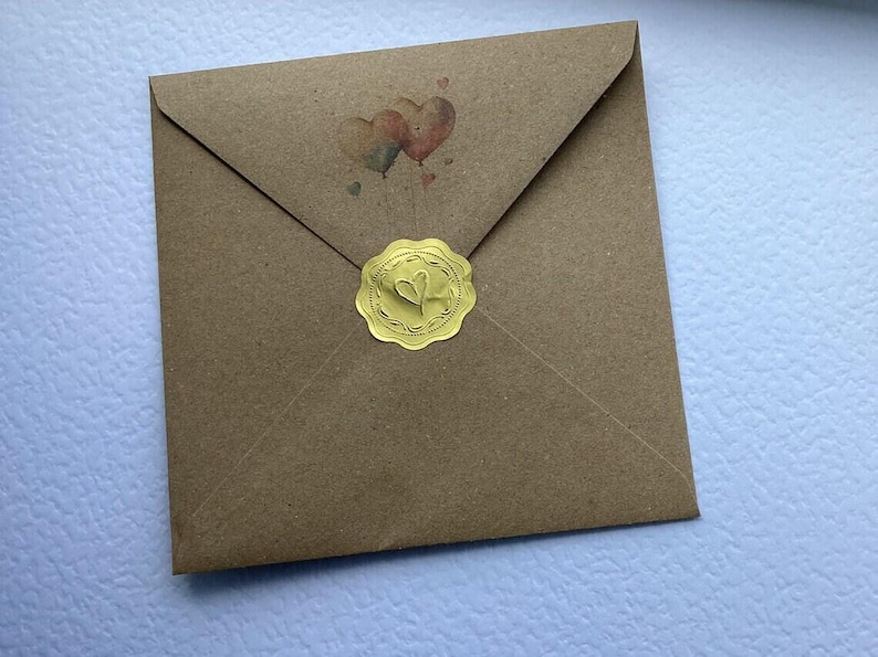 May include: A square brown kraft paper envelope featuring a small illustration of two heart-shaped balloons above the center. The envelope is sealed with a round, scalloped-edge gold foil sticker embossed with a central heart design.