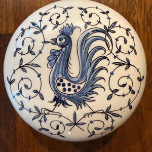 May include: A round white ceramic lid featuring a hand-painted blue rooster at the center. The rooster is surrounded by a delicate blue vine and leaf pattern that encircles the entire perimeter of the circular surface, creating a decorative border.