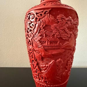 May include: A cinnabar red vase featuring intricate relief carvings of a landscape scene with traditional buildings, trees, and mountains. The surface is densely textured with detailed patterns, including a fluted base and a decorative neck.
