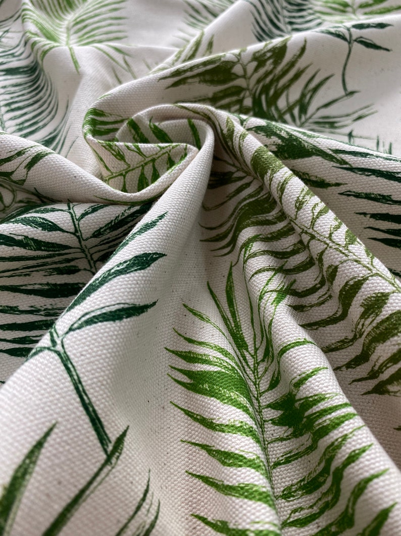 May include: White fabric with a green leafy pattern. The fabric is folded and shows the texture of the material.