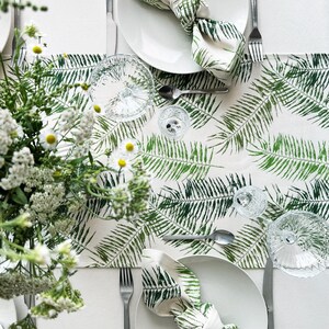 May include: A white tablecloth with a green fern pattern runner. Two white plates with green fern napkins are set on the runner. Silverware and clear glasses are also on the table.