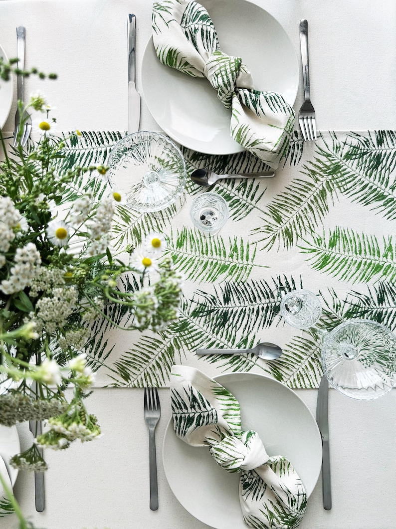 May include: A white tablecloth with a green fern pattern runner. Two white plates with green fern napkins are set on the runner. Silverware and clear glasses are also on the table.