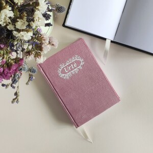 May include: Two notebooks, one with a pink cover and the word "Urte" in white lettering, and the other with a white cover and black spine.