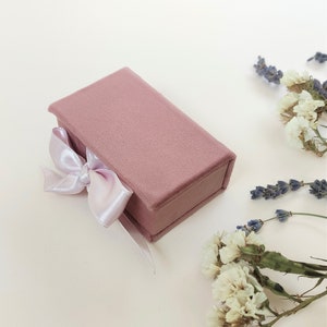 Può includere: Una piccola scatola di velluto rosa polveroso con un nastro di raso bianco legato in un fiocco. La scatola è su una superficie bianca.