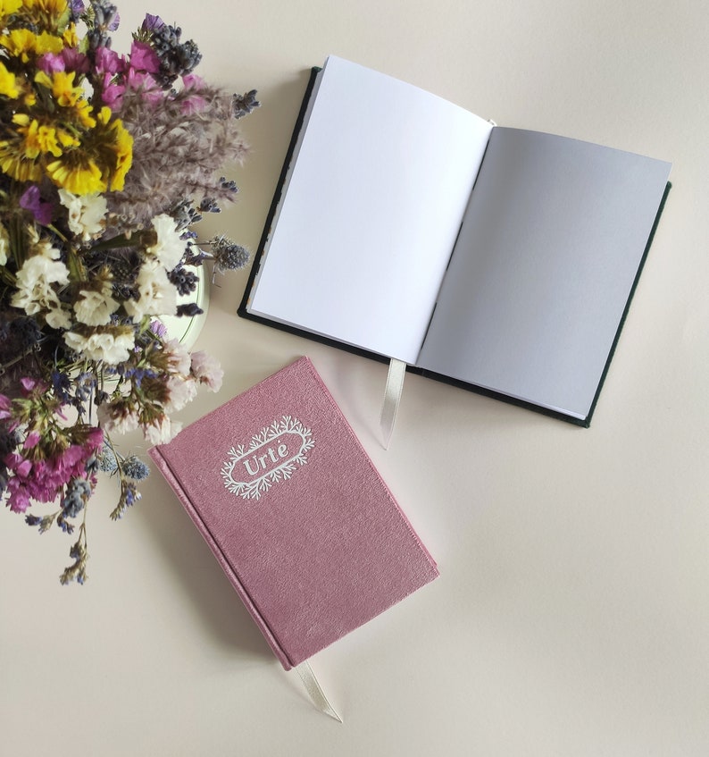 May include: Two notebooks, one with a pink cover and the word "Urte" in white lettering, and the other with a white cover and black spine.