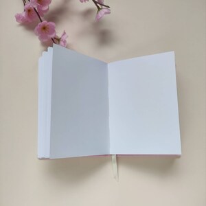 May include: A white blank notebook with a pink cover and a white ribbon bookmark. The notebook is open and lying flat on a light beige surface.