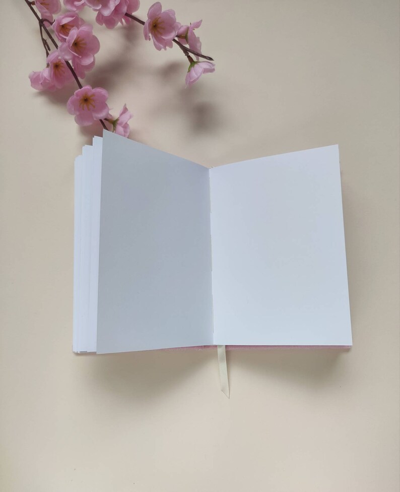 May include: A white blank notebook with a pink cover and a white ribbon bookmark. The notebook is open and lying flat on a light beige surface.