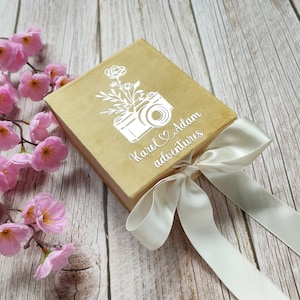 May include: A gold box with a white ribbon tied around it. The box has a white design of a camera with flowers and the text "Karol & Adam adventures".