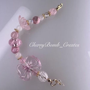 May include: A beaded charm featuring pink and clear beads, two translucent pink flower-shaped beads, and gold-toned metal spacers. The charm includes a lobster clasp at one end and a small ring at the other, with the text CherryBomb_Creates visible.
