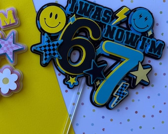 May include: Two birthday cake toppers with the text "I WAS NOW I'M" and the numbers "6" and "7". One is pink and blue, the other is black and blue. Both have smiley faces, stars, and lightning bolts.