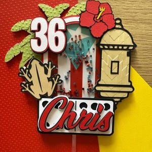 May include: A colorful cake topper with the number 36, a red hibiscus flower, and the name "Chris" in red script. The design includes a frog, palm leaves, and a lantern, all set against a red, white, and blue background.