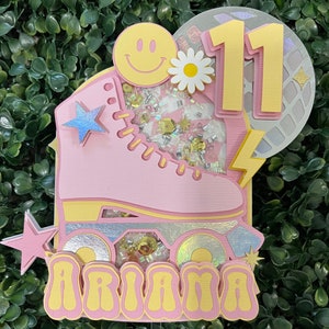 May include: Pink roller skate cake topper with a yellow smiley face, a white flower, and the number 11. The roller skate has glitter and a silver star. The name "Ariana" is written in yellow letters with pink outlines.