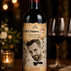 Puede incluir: Botella de vino tinto oscuro con una etiqueta personalizada que presenta un retrato en sepia de un hombre. La etiqueta dice "19 Crimes", "Happy Birthday, Love you loads" y "2026 Aaron". La botella est&aacute; sobre una superficie de madera.