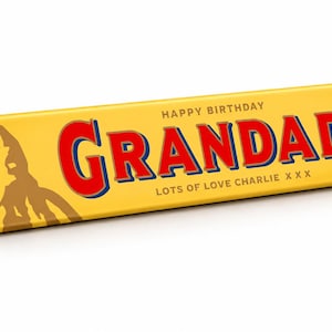 Pu&ograve; includere: Una barretta di cioccolato Toblerone triangolare in confezione gialla con il testo HAPPY BIRTHDAY GRANDAD LOTS OF LOVE CHARLIE X X X. Il lato mostra un grafico di montagna marrone e la data 18TH JUNE 2026 in un riquadro blu.